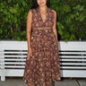 A woman standing in front of a fence with greenery  wearing a raisin floral tapestry midi caftan, belted and sleeveless. 