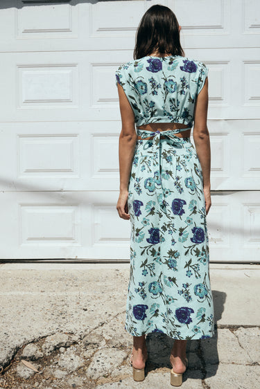 A woman standing outside by a white garage wearing an aqua ground floral tapestry skirt with a drop waist and side slits, paired with a matching cocoa top. 
Back view.