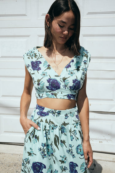 A woman standing outside by a white garage wearing an aqua ground floral tapestry skirt with a drop waist and side slits, paired with a matching cocoa top. Close up of waistline detail. 