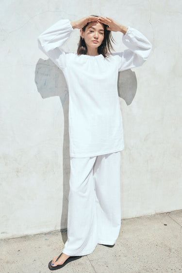 A woman wearing a white mini dress with long sleeves and a gathered neckline with matching trousers standing outside by a white wall. 