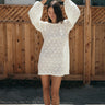 A woman standing outside by a brown wood fence wearing a lace mini dress with gold sandals.