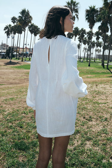A woman wearing a white mini dress with long sleeves and a gathered neckline, standing in an outdoor setting with palm trees in the background. Back View
