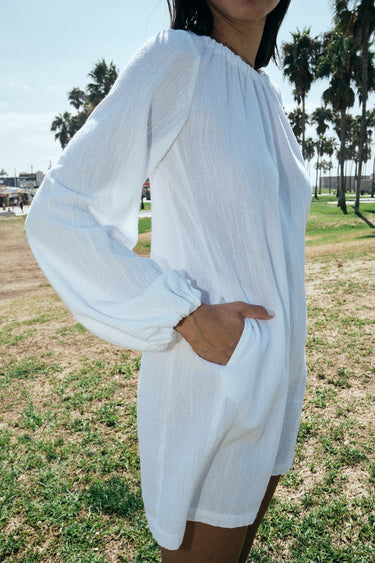 A woman wearing a white mini dress with long sleeves and a gathered neckline, standing in an outdoor setting with palm trees in the background.  Side View