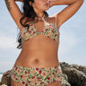 A woman on a beach by rocks wearing a beige ground with red, brown and green floral tapestry bikini bottom with a high rise and matching bikini top.