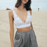 A woman standing on the beach wearing a white trimmed with cotton cluny lace  bra with adjustable straps and a drawstring back. Paired with salt and pepper woven trouser shorts. 