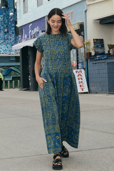 A woman at a beach boardwalk wearing a dark blue paisley tapestry maxi dress with pockets in the gathered skirt.