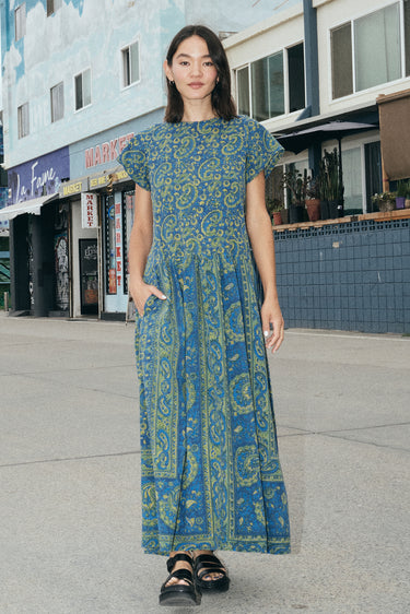 A woman at a beach boardwalk wearing a dark blue paisley tapestry maxi dress with pockets in the gathered skirt.