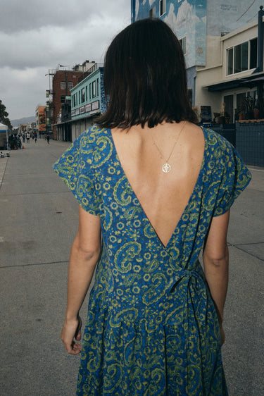 A woman at a beach boardwalk wearing a dark blue paisley tapestry maxi dress with pockets in the gathered skirt. Back view detail 