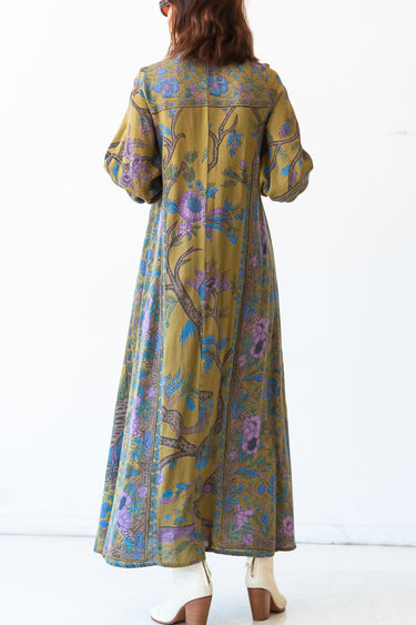 A woman standing in a studio with white walls wearing a maxi caftan cut in an olive green tree of life handloom tapestry, back view. 