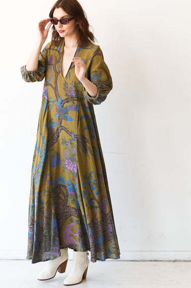 A woman standing in a studio with white walls wearing a maxi caftan cut in an olive green tree of life handloom tapestry.
