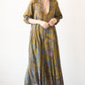 A woman standing in a studio with white walls wearing a maxi caftan cut in an olive green tree of life handloom tapestry.