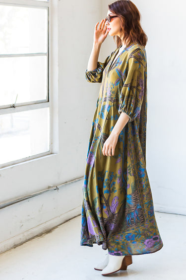 A woman standing in a studio with white walls wearing a maxi caftan cut in an olive green tree of life handloom tapestry. side view