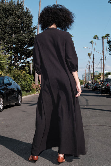 A woman standing outside e wearing a maxi length black v neck caftan, back view.