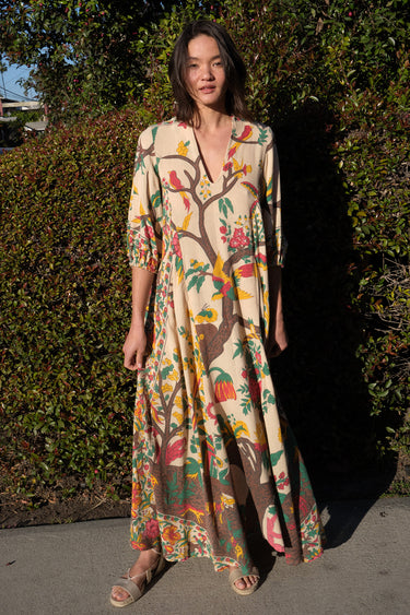 A woman standing outside by greenery wearing a maxi caftan cut in a tree of life handloom tapestry.