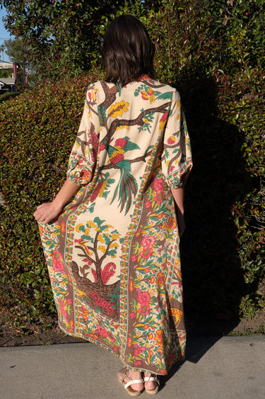 A woman standing outside by greenery wearing a maxi caftan cut in a tree of life handloom tapestry, back view.