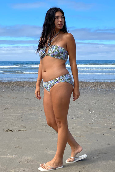 A woman on a beach wearing a white ground with lavender and blue floral tapestry bikini bottom with a low-rise design and matching bikini top.