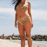 A woman on a beach wearing a beige ground with red, brown and green floral tapestry bikini bottom with a low-rise design and matching bikini top.