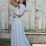 A woman standing outside by a mural wearing a white gauze with black stripe skirt with side slits and a drop waist, paired with a matching crop top. Side View