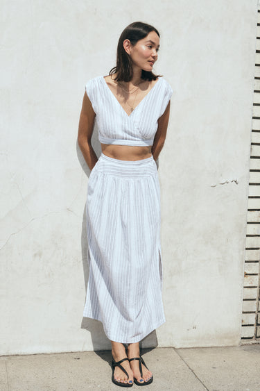 A woman standing outside wearing a white gauze stripe skirt with side slits and a drop waist, paired with a matching cocoa-colored top. 