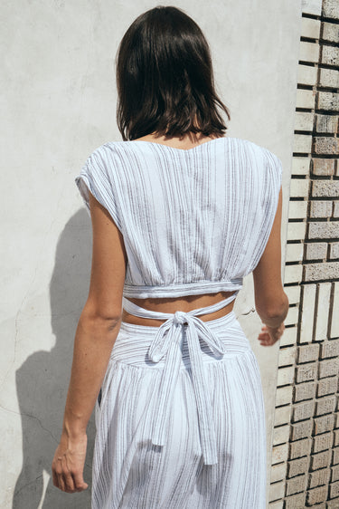 A woman standing outside in front of a white wall wearing a white with black stripe gauze crop top with matching skirt. Back View