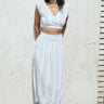 A woman leaning on a white wall, outside wearing a white with black stripe gauze crop top with matching skirt. 