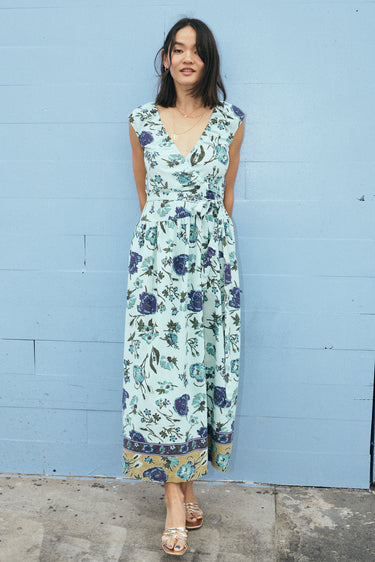 A woman standing by a light blue wall wearing a wrap dress cut in an aqua ground floral tapestry.
