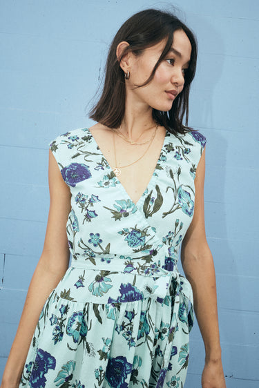 A woman standing by a light blue wall wearing a wrap dress cut in an aqua ground floral tapestry. Close up of top of dress. 