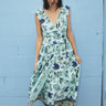 A woman standing by a light blue wall wearing a wrap dress cut in an aqua ground floral tapestry.