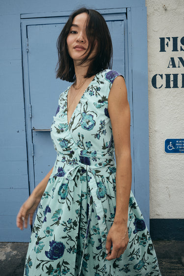 A woman standing by a light blue door wearing a wrap dress cut in an aqua ground floral tapestry.