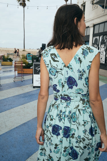 A woman standing at the beach wearing a wrap dress cut in an aqua ground floral tapestry, back view.