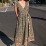 A woman standing on the sidewalk wearing a green floral tapestry midi dress with an elastic neckline and pockets.