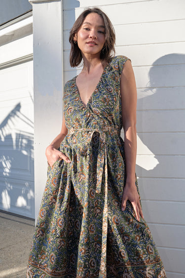 A woman standing on the sidewalk wearing a green floral tapestry midi dress with an elastic neckline and pockets.