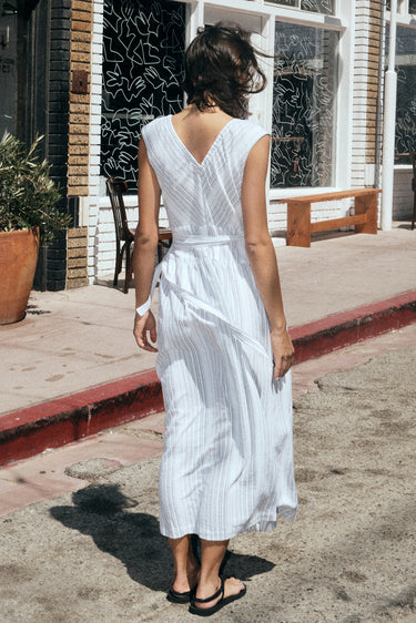 A woman standing in a street with shops wearing a long white with black stripe gauze sleeveless wrap dress, back view.