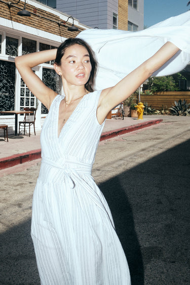 A woman standing in a street with shops wearing a long white with black stripe gauze sleeveless wrap dress while putting on a white jacket.