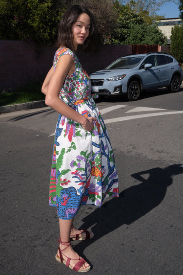 A woman standing on the sidewalk wearing a floral tapestry midi dress with an elastic neckline and pockets.