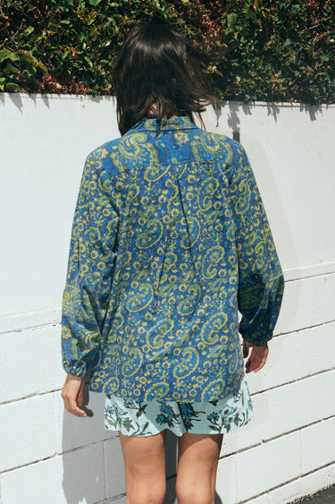 A woman standing outside by a wall wearing a shirt jacket cut from a dark blue paisley tapestry, with an aqua tapestry short, back view.