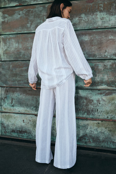 A woman outside by a verdigris wall wearing a long sleeved white with black stripe gauze shirt jacket with a matching trouser, back view.