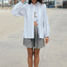 A woman on a beach side walk wearing a long sleeved white with black stripe gauze shirt jacket  paired with a gray short and a white bra top. 