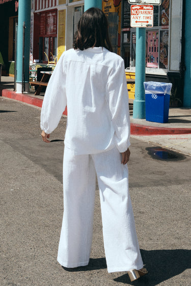 A woman standing at the beach boardwalk wearing a white osnaburg shirt jacket with patch pockets and a matching trouser. Back View