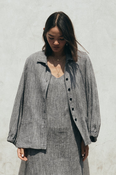 A gray unlined jacket with big patch pockets and an elastic shirred sleeve cuff, worn by a model facing away from the camera. She is wearing the jacket with a matching sheath dress. Close up detail. 