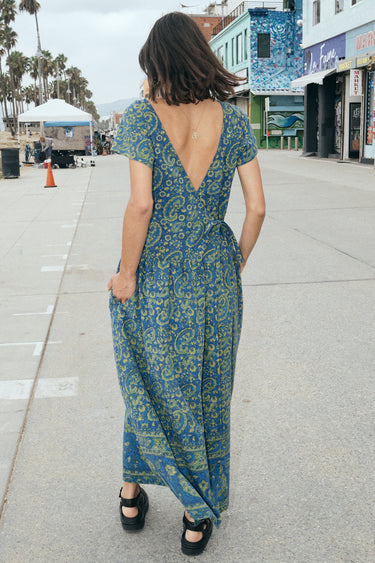 A woman at a beach boardwalk wearing a dark blue paisley tapestry maxi dress with pockets in the gathered skirt. Back View
