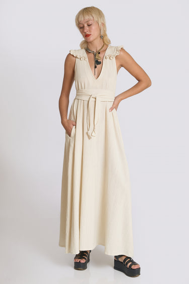 A woman standing  in a photo studio with white walls wearing an off white alabaster circular flared sleeveless maxi dress with ruffles around the yoke, with an obi belt.  