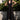 A woman standing outside in front of a lime tree wearing a black circular flared sleeveless midi dress with ruffles around the yoke.  