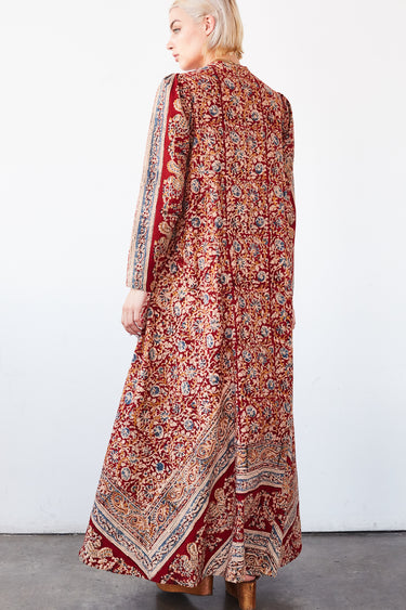 A woman in a studio with white walls wearing a red with blue flowers tapestry caftan with long sleeves. Back View