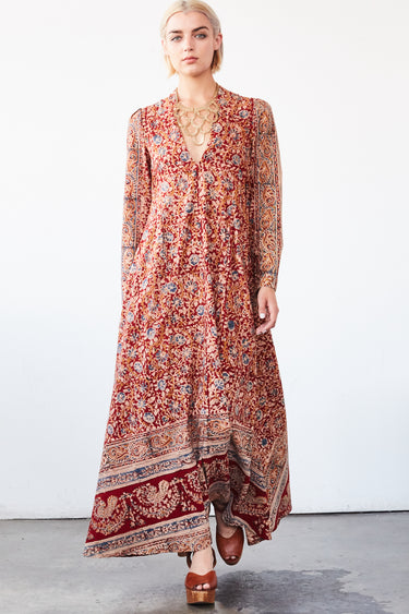 A woman in a studio with white walls wearing a red with blue flowers tapestry caftan with long sleeves. 