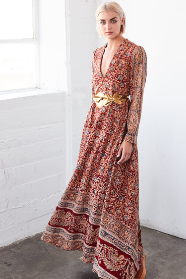 A woman in a studio with white walls wearing a red with blue flowers tapestry caftan with long sleeves. She is wearing a gold belt. 