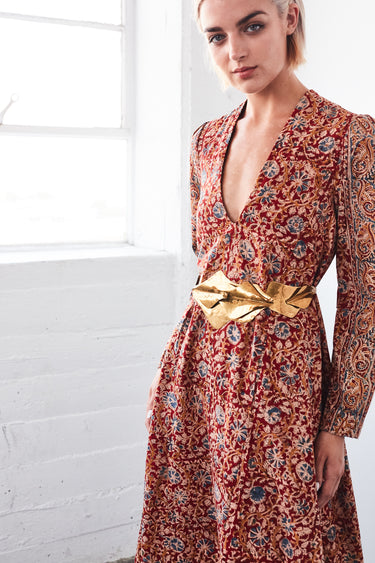 A woman in a studio with white walls wearing a red with blue flowers tapestry caftan with long sleeves.  Close up, with gold belt. 