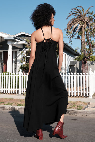 A model wearing a black circular maxi slip dress standing outside near a white picket fence with a house in the background. Back View