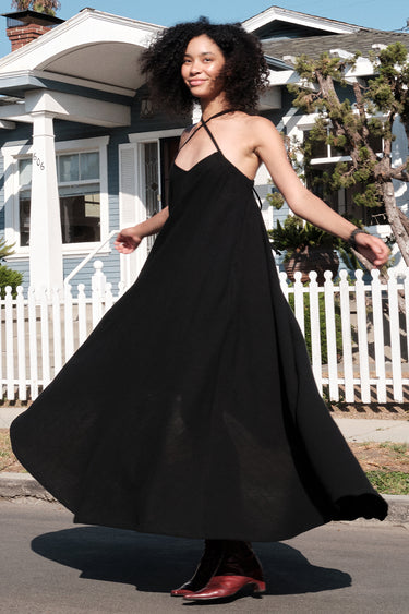 A model wearing a black circular maxi slip dress standing outside near a white picket fence with a house in the background.