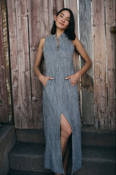 A model wearing a gray maxi shift dress with a front zipper and pockets, standing near a wooden fence.